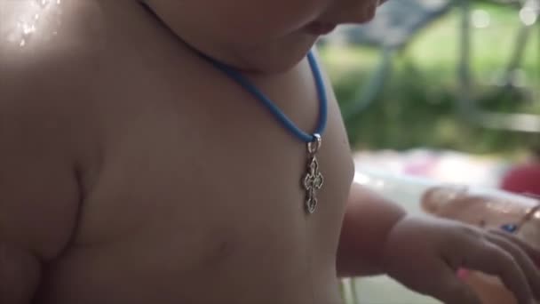 在外面浴缸里洗澡的婴儿的身体 脖子上有一个基督教十字架的特写 蓝色的绳子水滴散落 阳光灿烂 洗礼后 — 图库视频影像