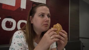 Aç gözlü genç bir kadın bir fast food restoranında et burger yiyor. Sandviçi paylaşmayı teklif etti. Mutlu, gülümseyen bir kadının portresi yemek yiyor ve kameraya bakıyor, konuşuyor. Yüzük ve