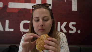 Aç gözlü genç bir kadın bir fast food restoranında et burger yiyor. Yemek yiyen ve kameraya bakan kızgın bir kadının portresi..