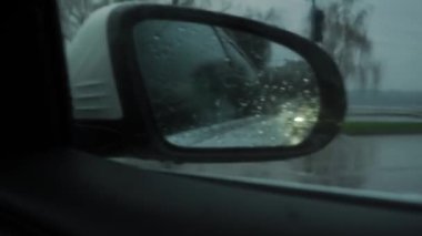 Side mirror of a car driving through the city in a downpour, in winter, autumn, spring, in cloudy weather. Close-up on drops on the window. Mobile camera. In the reflection of cars on the road