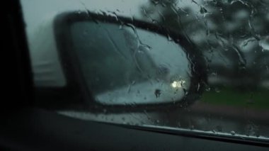 Side mirror of a car driving through the city in a downpour, in winter, autumn, spring, in cloudy weather. Close-up on drops on the window. Mobile camera. In the reflection of cars on the road