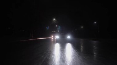 a white car slowly drives along the highway due to bad weather, it is raining, headlights shine with white fire, wet slippery asphalt. big truck driving behind