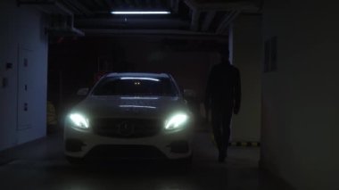 Ukrayna, Zaporozhye - 25 Aralık 2022: Yeraltında yağmurdan düşen damlalarla kaplı beyaz bir araba var, farlar açık, adam yavaşça yaklaşıyor, kapıyı açıyor ve orada oturuyor.