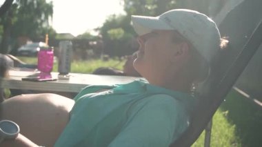 Yeşil mayosu ve şapkası olan hamile bir kadın, güneşleniyor, kır evinde güneşleniyor. Yazın, ağaçların arkasında, güneşin önünde.