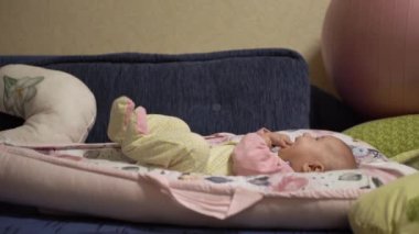 Neşeli küçük bir bebek, ellerini ve parmaklarını ağzına koyar, seğirir, bacaklarını oynatır, oynar, eğlenir, odayı inceler. Bebek tulumu giymiş bir çocuk, mavi bir kanepede kozanın içinde. Yan görünüm