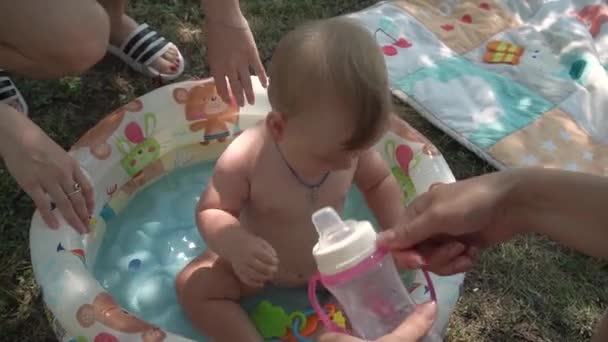 母親は暖かい澄んだ水のプールに座っている間に小さな女の子にベビーカップから飲み物を与えます おもちゃが浮かぶ子供の首に青いレース — ストック動画