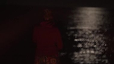 Ay 'ın yansıması ve suyun üzerindeki ay ışığıyla gece okyanusu. Yalnızlık. Kapüşonlu bir kız karanlık denize, kamp alanındaki sahile bakıyor. Bokeh 'te ay ışığı