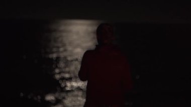 Ay 'ın yansıması ve suyun üzerindeki ay ışığıyla gece okyanusu. Yalnızlık. Kapüşonlu bir kız karanlık denize, kamp alanındaki sahile bakıyor. Bokeh 'te ay ışığı
