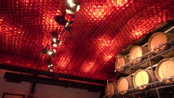 ワイナリー ワインバー 明るいランプは赤いガラスびんから成っている天井で 壁に対して 巨大な木製樽の完全なワイン リキュール デカンターに注ぐためのパイプが付いている棚があります — ストック動画