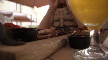 Bir çift yaz terasında bir restoranda öğle yemeği yiyor, sırayla tatlı sosa sebze bandırıyor, ahşap bir tahtanın üzerinde kabuklu aromalı domuz pirzolası yiyor, ve yanında bir bardak bira duruyor.