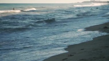 Küçük deniz dalgası, bulanık yumuşak köpüklü dalgalar altın kumlu sahil şafağını yıkıyor. Yürüyen insanlar, su kayakları kumsalları, mercanlar, Grizon tekneleri. Tatil eğlencesi konsepti. Soyut deniz yaz okyanusu gün batımı