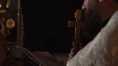 Uzun siyah sakallı, beyaz cüppeli bir rahip elinde İncil 'i tutuyor ve dua okuyor. Öte yandan, büyük bir altın Ortodoks haçı tutuyor. Alacakaranlıkta çekim yapıyor.
