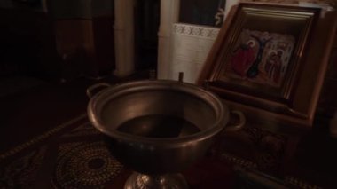 Masanın üzerinde kutsal su bulunan büyük gümüş bir kase, İncil 'in eski bir kitabı ve altın çerçeveli büyük bir ikonun yanında, bir kilisede çekim yapan alacakaranlık ve sessizlik var.