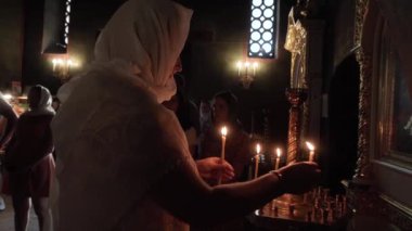 Kafasında hafif bir eşarp ve elinde bir bilezik olan bir kadın, Ortodoks kilisesinde bir cenaze masasının üzerine mum yakıyor, her yerde alacakaranlık var, her şey altın, insanlar...
