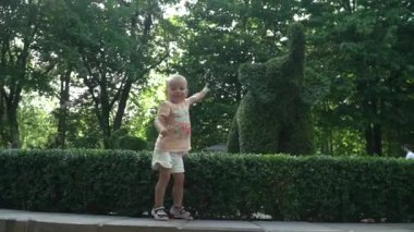 Küçük bir çocuk, kız, çalılıktan fil figürü, peyzaj ile park. Bagajında ve burnunda. Güler, gülümser, zıplar. Şortlu, tişörtlü, sandaletli, kısa saçlı. Gün, yaz