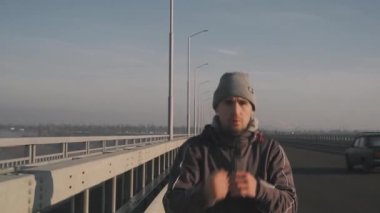 Sıcak giysiler içinde genç bir atlet sabah yürüyüşüne çıktı. Yol köprüsü boyunca, arabalar sürüyor. Sabah sisi arka planda görülebiliyor.