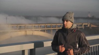 Sıcak giysiler içinde bir maraton koşucusu ve boş bir yolda güvenli ve odaklı bir şekilde koşan bir şapka, arka planda bir yol köprüsü görebilirsiniz, sabah rüzgarlı sisi, güneş parlıyor, soğuk