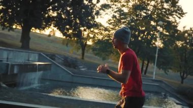 Kırmızı tişörtlü ve şapkalı bir atlet merdivenlerden yukarı koşuyor, özgüvenli hareketler, konsantrasyon, hızlı el hareketleri, dekoratif bir şelale ve gün batımına karşı.