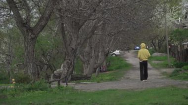 Sarı kapüşonlu bir koşucu, yemyeşil ağaçlarla çevrili manzaralı bir patikada hızla koşar.