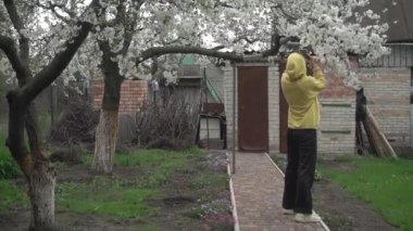 Zarif sarı kapüşonlu ve spor ayakkabılı kız çiçek açan ağacın güzelliğinden hoşlanıyor, kasvetli bir yaz gününde kır evinin bahçesinde video ve fotoğraf çekiyor.