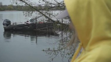 Sarı kapüşonlu bir kız ve kumlu bir nehir kıyısında sakince ve barışçıl bir şekilde doğanın tadını çıkaran bir şapka. Arka planda bir adamla birlikte küçük bir motorlu tekne demirli.