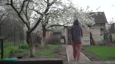 Eşofmanlı genç bir kız kapıya doğru yürüyor, kiraz ağaçları çiçek açıyor, çimenler yeşil.