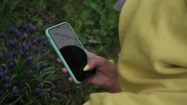 Parlak manikürlü sarı kapüşonlu bir kızın arkasından modern bir akıllı telefondan şirin mor çiçeklerin şık fotoğraflarını çekiyor.