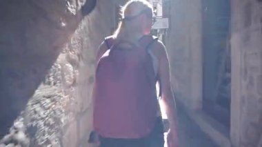 Kamera, genç bir turisti takip ediyor, sarışın, güneş gözlüğü takan, pembe bir sırt çantası ve şortu, eski şehrin dar sokaklarını keşfedip etrafa bakıyor. İyi günler. Yaz