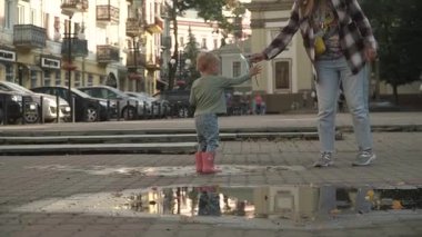 Kot pantolonlu ve uzun saçlı bir kız su birikintisindeki lastik çizmeli küçük bir kıza yel değirmeni oyuncağı veriyor, neşeyle suya indiriyor, neşe, yavaş çekim, serin yaz günü