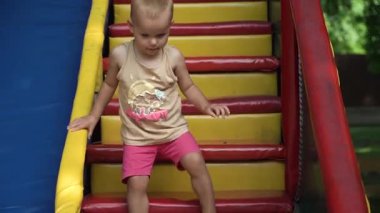Sarı saçlı, pembe şortlu ve tişörtlü bir kız hızla ve dikkatle tırabzanlara tutunarak çok renkli basamakları tırmanır. Başka bir çocuğa yol vermek, sıcak bir yaz günü, neşe, ilgi, merak
