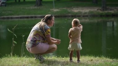 Kız dikkatle izliyor, bir çocuğun yanında oturuyor, parmağını sudaki kuşlara ve balıklara doğrultuyor, yazın Central City parkında manzaranın ve doğal yeşil gölün tadını çıkarıyor.