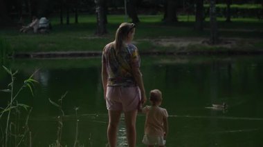 Yazlık elbiseli genç bir anne Central Park 'taki göl kenarındaki çimen ve ağaçlarla dolu yeşil bir bankta el ele dururken kuşlara, balıklara, balıkçılara ve çocuklara bakıyor.