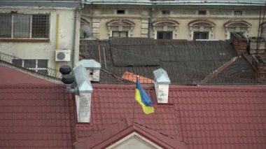 Mavi ve sarı bir Ukrayna bayrağı rüzgarda dalgalanır kafatasına benzer çatısı olan eski bir şehir, arkasında eski mimari pencereleri ve verandaları kırık, bakımsız bir bina. İvano-Frankivsk