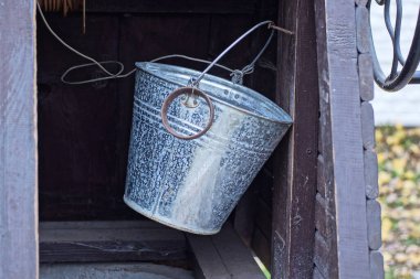 Bir gri metal kova, dışarıdaki eski bir kırsal kuyunun içindeki kahverengi ahşap duvarda asılı.