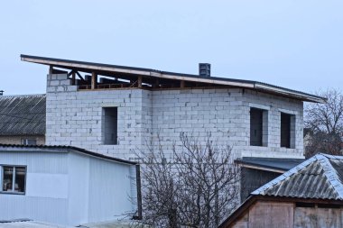  Tamamlanmamış beyaz tuğlalı bir evin parçası. Çatısında tahtalar ve gri gökyüzüne karşı ahşap kahverengi şekiller var.