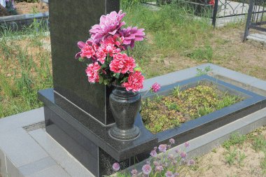 Mezarlıktaki bir mezarın üzerindeki granit bir anıtta kırmızı sanatsal çiçekli siyah bir mermer vazo.