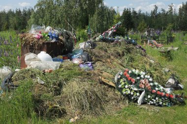 Kuru çimenlerde paslı kahverengi çöp tenekesi ve mezarlığın yakınındaki sokakta eski cenaze çelenkleri olan bir çöp yığını.