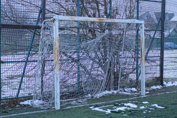 鉄の柵の近くの白い雪の中の緑の芝生冬の通りの白い雪の中のサッカー場の近くに引き裂かれたメッシュを持つ古い金属錆びた門 — ストック写真