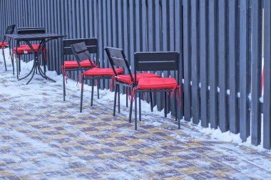 Kırmızı koltuklu siyah bir sıra beyaz karda kaldırımda duruyor. Kış sokağındaki ahşap bir çit duvarına karşı.