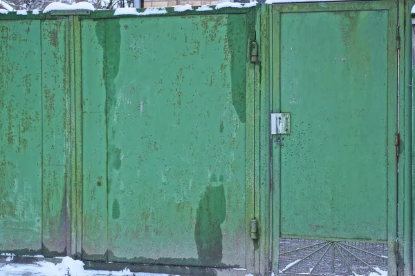 wet green metal gate and closed iron door in white snow on a rural street