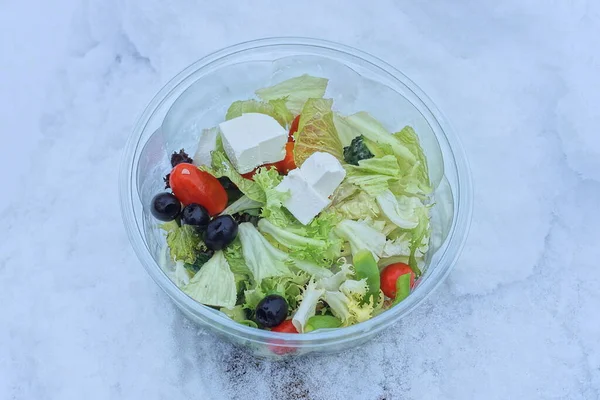 Greek salad with olives and cheese in a plastic plate stands on white snow on the street