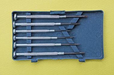 Sarı bir masanın üzerindeki plastik bir kutuda gri siyah metal tornavidalar.