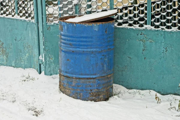one old blue metal barrel stands in a drift of white snow against a green fence wall on a winter street
