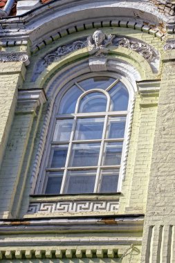 Yeşil tuğlalı bir duvarda büyük, eski, beyaz bir pencere. Üzerinde fresk ve sokaktaki tarihi bir binanın desenleri var.