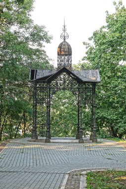 one open black metal gazebo with a wrought iron pattern on the gray sidewalk of an alley among green vegetation in a summer park
