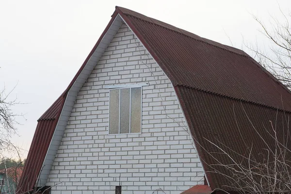 one attic of a white brick private house with a window under a brown roof against a gray sky and tree branches