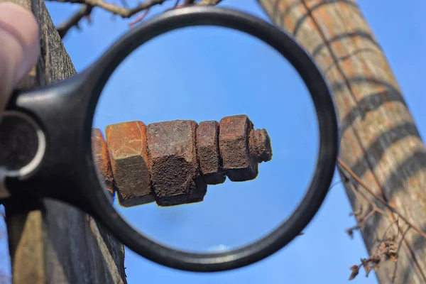 magnifying glass in hand magnifies a long iron bolt with brown rusty nuts on a gray wooden structure outdoors against a blue sky