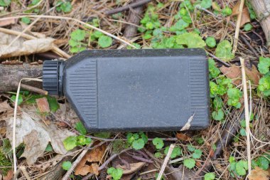 Eski bir siyah plastik kutunun çöpü gri toprakta ve sokaktaki yeşil bitki örtüsünde yatıyor.