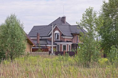 Siyah kiremitli bir çatının altında yeşil çimlerde ve bitkilerle kaplı bir çitin arkasında gri bir gökyüzüne karşı büyük kahverengi bir ev.