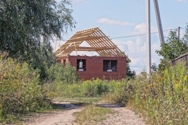 Kırsal bir sokakta mavi gökyüzüne karşı yeşil bitki örtüsü içinde ahşap çerçeveli, tamamlanmamış kırmızı tuğlalı bir ev.